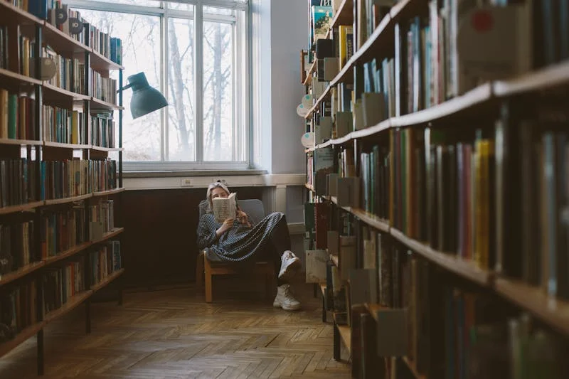 Fotografía de una mujer estudiante en una biblioteca sentada junto a su libro donde la luz es un poco obscura