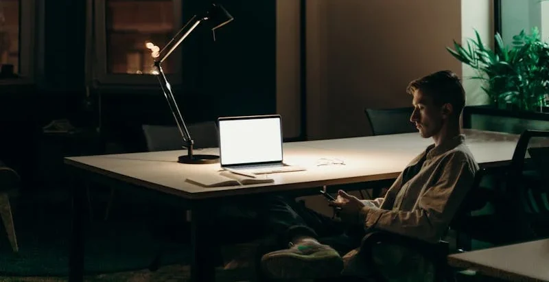 Fotografía de un estudiante en una oficina un poco obscura con una lampara de fondo, este estudiante revisa su computadora.